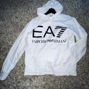 Vit hoodie från Emporio Armani EA7 - Vit hoodie från Emporio Armani EA7 med stor svart logga framtill och mindre logga baktill. Tröjan har huva och långa ärmar, perfekt för en stilren och sportig look.