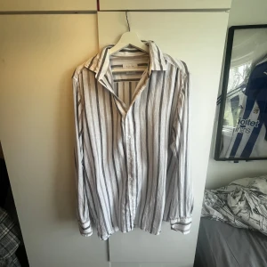 Randig skjorta från Massimo Dutti - Säljer en snygg vit skjorta med gråa och beige ränder från Massimo Dutti. Skjortan har klassisk krage, knäppning framtill och långa ärmar. Perfekt för dig som gillar en stilren och avslappnad look.