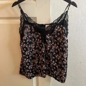 Svart blommig linnetopp med spets från H&M - Säljer en svart linnetopp från H&M med små rosa och vita blommor. Toppen har tunna axelband och vacker spetsdetalj längs urringningen. Perfekt för en somrig look och lätt att matcha med jeans eller kjol.