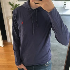 Blå hoodie från Polo Ralph Lauren - Säljer en blå hoodie från Polo Ralph Lauren med röd broderad logga på bröstet. Tröjan har långa ärmar, huva med dragsko och är tillverkad i mjuk bomull. Perfekt för en avslappnad stil. Storlek S