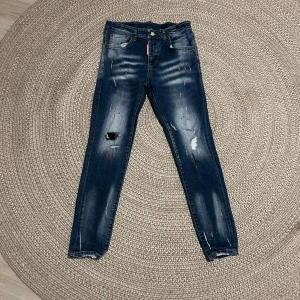 Mörkblå slitna jeans med färgstänk - Snygga mörkblå jeans med slitningar och färgstänk på benen. Klassisk femficksmodell med raka ben och normal passform. Jeansen har en något tvättad look och detaljerade slitningar på låren och knäna.