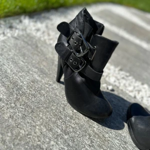 Svarta boots med spännen och klack - Säljer ett par svarta boots med hög klack och två breda spännen i silver. Skorna har rund tå och är tillverkade i ett material som ser ut som skinn. Perfekta för dig som vill ha en edgy stil.