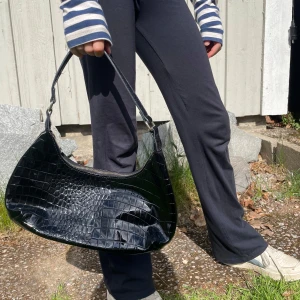 Svart axelväska i mönstrat skinn - Snygg svart axelväska i mönstrat skinn. Väskan har en böjd form och stängs med dragkedja upptill. Perfekt storlek för det viktigaste och ger en trendig touch till din outfit. 