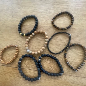Bruna o svarta armband - Säljer ett set med åtta armband i olika färger och material. Armbanden är gjorda av svarta och bruna pärlor i trä och sten, vissa med lavasten och andra med slät finish. Perfekta för att mixa och matcha till din stil.