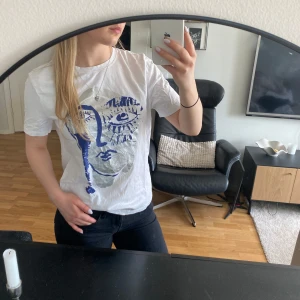 Vit t-shirt med konstnärligt tryck - Vit t-shirt med ett stort konstnärligt tryck i blått och silver på framsidan. T-shirten har rund hals och korta ärmar, perfekt för en avslappnad stil.
