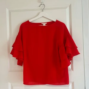 Röd blus med volangärmar från H&M - Säljer en röd blus från H&M med bred rund halsringning och volangdetaljer på ärmarna. Blusen har en lös passform och är tillverkad i ett lätt, syntetiskt material. Perfekt för dig som vill ha något färgstarkt och stilrent.PRIS GÅR ATT DISKUTERAS