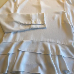 Vit volangblus från Zara Woman - Säljer en stilren vit blus från Zara Woman med långa ärmar och dubbla volangdetaljer nedtill. Blusen har knappar vid ärmsluten och är tillverkad i ett mjukt, lätt material. Perfekt för en trendig och elegant look.