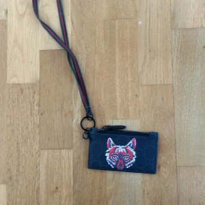 Svart plånbok med vargmotiv från Gucci - Säljer en svart plånbok från Gucci med broderat vargmotiv på framsidan. Plånboken har dragkedja och avtagbart axelband i rött och grönt. Perfekt för att bära det viktigaste med stil. Hör av er för fler frågor eller funderingar 👍🏼