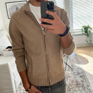 Beige stickad kofta med dragkedja - Säljer en beige stickad kofta med dragkedja och ribbade muddar. Perfekt för lager-på-lager och enkel att matcha med olika outfits. Klassisk och stilren design med hög krage.