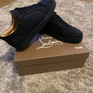 Svarta sneakers från Christian Louboutin - Säljer ett par svarta sneakers från Christian Louboutin med klassisk röd sula och diskret broderad logga på hälen. Skorna har snörning och är tillverkade i mocka med svarta detaljer och rund tå.
