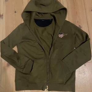 grön hoodie från GANT med dragkedja - Olivgrön hoodie från GANT med dragkedja framtill och broderad logga på bröstet. Mjuk bomullskänsla, perfekt för en avslappnad stil. Har en röd fläck vid märket, de från en bläck penna så jag vet inte om de går bort eller inte, därför de låga priset! 