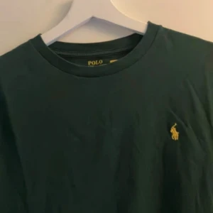 Mörkgrön t-shirt från Polo Ralph Lauren - Snygg grön t-shirt från Ralph Lauren med en liten gul broderad logga på bröstet. Perfekt för en stilren look. Storlek är 18y men passar ca 170-180 cm. Nypris ca 650kr
