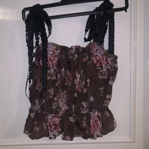 Brun blommig linnetopp från Brock Collection x H&M - Supersöt brun linnetopp från Brock Collection x H&M med blommigt mönster i rosa och lila toner. Toppen har volang nertill och breda axelband i svart. Perfekt för sommardagar och ger en romantisk vibe.