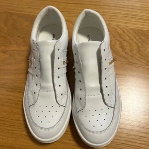 Vita sneakers med gulddetaljer från Sofie Schnoor - Säljer ett par vita sneakers från Sofie Schnoor med guldfärgade nitar på sidan. Skorna har perforerade detaljer upptill och en klassisk rund tå. Perfekta för dig som gillar stilrena och trendiga skor med en twist. Endast använda fåtal gånger! Skosnören saknas.