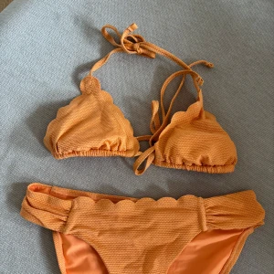 Orange bikini med vågig kant - Säljer en orange bikini med vågiga kanter och ribbad struktur. Överdelen har trekantsmodell med knyt i nacken och ryggen.  Storlek S på både över och underdel. Använd 1 gång i mycket gott skick!