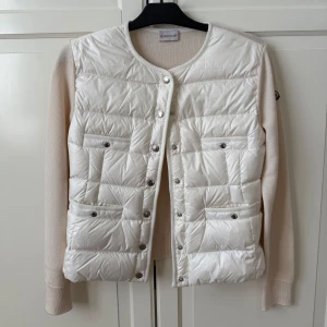 Vit cardigan med stickade ärmar från Moncler - Säljer en snygg vit cardigan från Moncler med stickade ärmar i vit. Västen har rund halsringning, tryckknappar framtill och två fickor med knapp. Moncler-logga på ärmen. Perfekt för lager-på-lager och stilren look. Storlek S! 