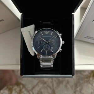 Emporio Armani herrklocka med blå urtavla - Snygg herrklocka från Emporio Armani med blå urtavla och silverfärgad länk i metall. Klockan har tre subdials, datumvisning och tydliga indexmarkeringar. Perfekt accessoar för dig som gillar stilrena detaljer.Den här helt ny med kvitto och kostar igentligen 2399kr