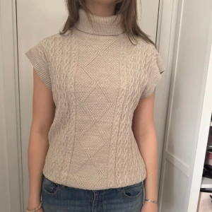 Beige stickad polotopp med korta ärmar - Säljer en beige stickad polotopp med korta ärmar och kabelstickat mönster framtill. Tröjan har hög polokrage och är perfekt för lager-på-lager. Snygg till både jeans och kjol!