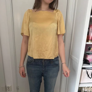 Gul blus med kort ärm - Säljer en söt gul blus med fyrkantig ringning och korta, luftiga ärmar. Blusen har en lätt glansig yta och är tillverkad i ett tunt, mjukt material. Perfekt för varmare dagar och enkel att matcha med jeans eller kjol.