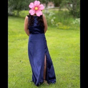 Marinblå långklänning med slits - Marinblå långklänning i satin med slits framtill och vacker drapering i ryggen. Klänningen har ärmlös design och en stilren, figurnära passform som passar perfekt till fest eller bal. Priset är inte hugget i sten