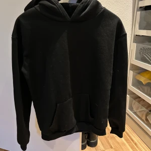 Svart hoodie med ficka - En klassisk svart hoodie med huva och magficka. Tröjan har långa ärmar och ribbade muddar vid ärmslut och nederkant. Perfekt för en avslappnad stil.