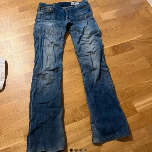 Blå lågmidjade bootcut jeans från Levis 527 - Lågmidjade blå jeans från Levis modell 527 med bootcut passform. Jeansen har fem fickor, dragkedja och knapp i midjan samt tydliga slitningar för en cool look. Perfekta för dig som gillar en avslappnad stil. Vintage och jättesnyggt!