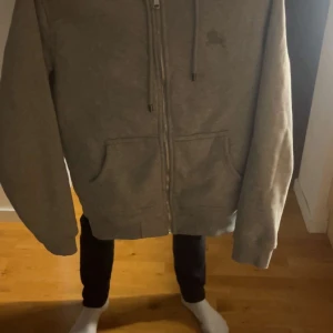 Grå zip-hoodie från Burberry - Säljer en grå hoodie med dragkedja från Burberry. Utsidan är enkel och stilren, medan insidan har det klassiska rutiga Burberry-mönstret i beige, svart och rött. Två fickor framtill och justerbar huva med snörning.
