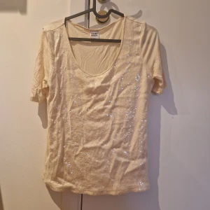 Vit glittrig t-shirt från Vero Moda - Säljer en vit t-shirt från Vero Moda med glittriga detaljer framtill. T-shirten har korta ärmar och en djup rundringning. Perfekt för dig som vill ha något enkelt men ändå med lite extra bling.