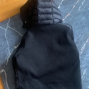 Svart jacka med quiltad huva - Säljer en svart jacka med quiltad huva och ribbad struktur på kroppen. Jackan har dragkedja och en sportig look, perfekt för dig som gillar stilrena plagg med lite edge.