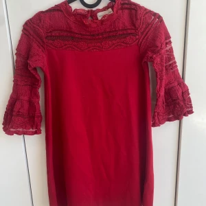 Röd klänning med spets och volangärmar - Säljer en röd klänning med vackra spetsdetaljer upptill och på ärmarna. Klänningen har trekvartsärmar med volanger och en rak passform. Perfekt för dig som vill sticka ut med en färgstark look. Klänning är för barn runt 6års åldern
