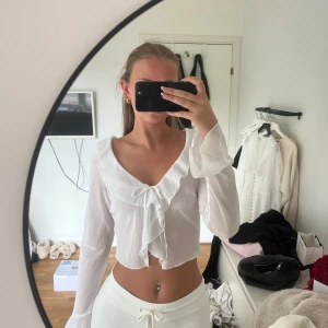 Vit blus med volang och knyt - Säljer en söt vit blus med volangkant och knyt framtill. Blusen har långa ärmar och en djup v-ringning. Perfekt för dig som gillar romantisk och feminin stil. Även perfekt att styla till en ljus kjol i sommar. 