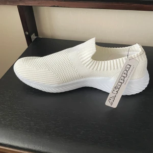 Vita slip-on sneakers från Kappa - Säljer ett par vita stickade slip-on sneakers från Kappa. Skorna har en ribbad kant runt öppningen och en platt, vit sula. Perfekta för dig som gillar enkel och sportig stil. Märkeslogga på sidan och på innersulan.