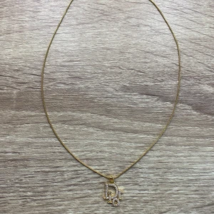 Stainless guldhalsband med Dior-hänge - Elegant stainless halsband i guld med ett gnistrande Dior-hänge dekorerat med små stenar. Kedjan är tunn och smidig med ett klassiskt lås. Går att ducha med den med. Detta är perfekt för dig som vill ha något stilrent men ändå iögonfallande.