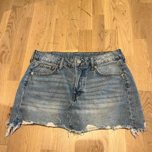 Jeanskjol från &Denim - Säljer en klassisk blå jeanskjol från &Denim i modell med råa, fransiga kanter och slitningar. Kjolen har fem fickor, bälteshällor och knappgylf. Perfekt för en avslappnad stil.                 Priset kan diskuteras 