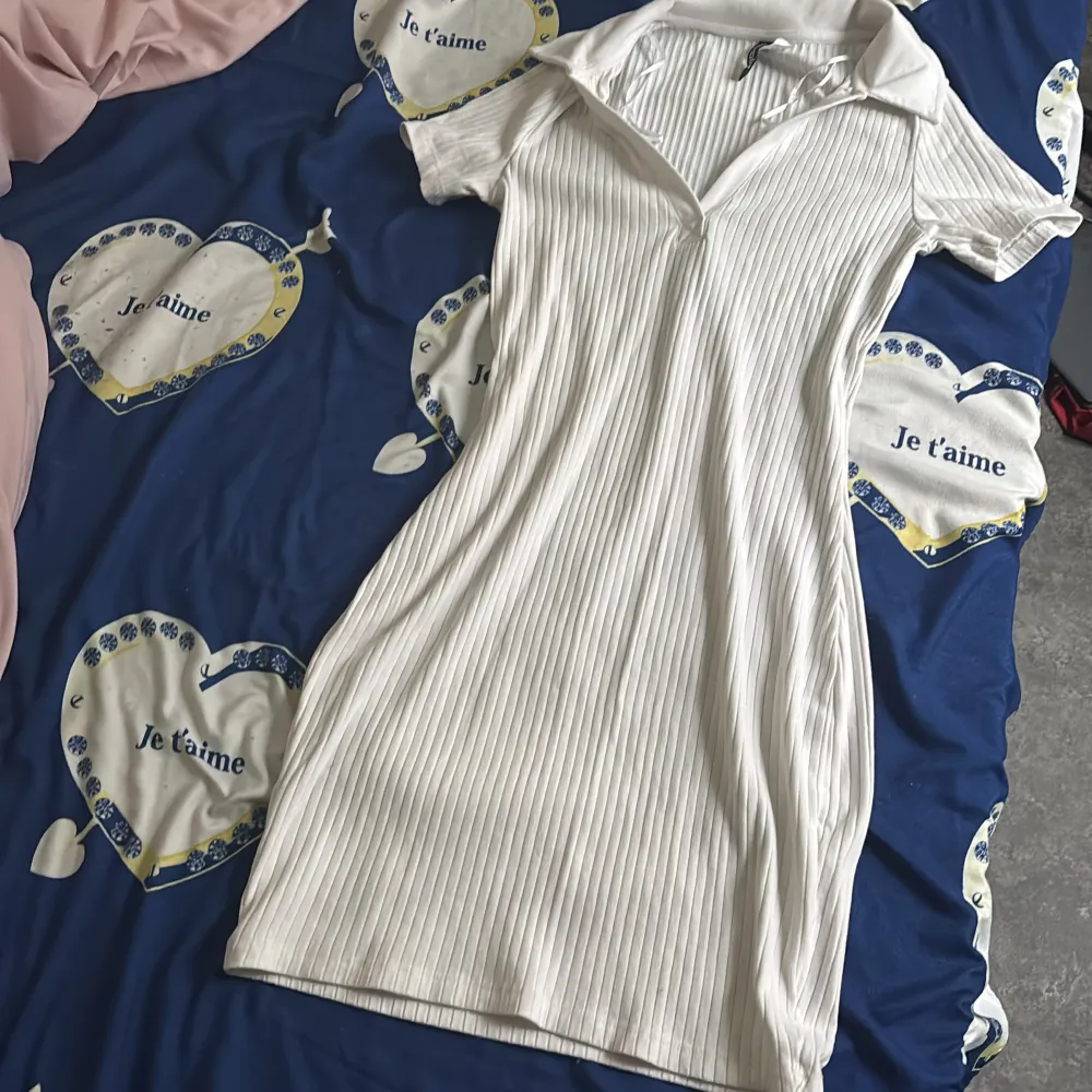 Säljer en vit ribbad kortklänning med krage och v-ringning. Klänningen har korta ärmar och en figurnära passform som framhäver siluetten. Perfekt för en fräsch och stilren look.. Mekot.