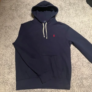 Ralph Lauren hoodie - Säljer en klassisk mörkblå hoodie från Polo Ralph Lauren med vit dragsko och röd broderad logga på bröstet. Tröjan har känguruficka och mjuk insida, perfekt för en avslappnad stil.