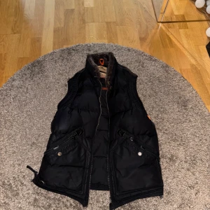 Svart dunväst med dragkedja - Snygg svart dunväst med hög päls krage och dragkedja framtill. Västen har två snedställda fickor med knappar och en sportig look. Perfekt att slänga över en hoodie eller tröja för extra värme.