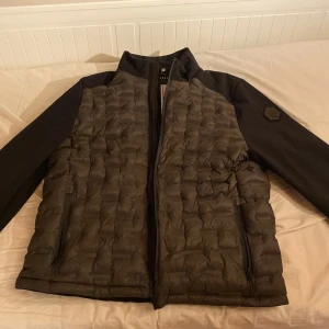 Svart pufferjacka från SMOG - Säljer en svart pufferjacka från SMOG i storlek L. Jackan har quiltad framsida, släta svarta ärmar och två dragkedjefickor framtill. Perfekt för kyligare dagar och har en stilren look med hög krage.