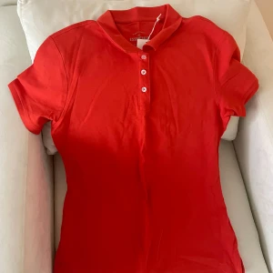 Röd pikétröja från The Essentials by Race Marine - Säljer en klassisk röd pikétröja från The Essentials by Race Marine. Tröjan är i bomull, har korta ärmar och tre vita knappar framtill. Perfekt för en stilren och sportig look.