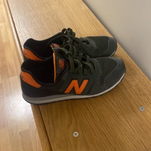 New Balance sneakers i grönt och orange - Säljer ett par New Balance sneakers i grönt med orange detaljer och logga. Skorna har snörning och en klassisk sportig design med rund tå och platt sula. Perfekta för dig som gillar färg och vill ha bekväma skor till vardagen.