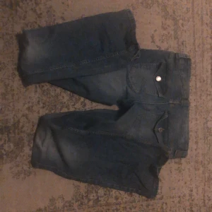 Blå bootcut jeans med fickdetaljer - Säljer ett par klassiska blå jeans med bootcut passform. Byxorna har snygga fickor med lock och knapp bak, samt lågmidjad  midja. Perfekta för dig som gillar en tidlös jeansstil med lite utsvängda ben.