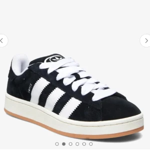 Adidas Campus  - Klassiska Adidas Campus sneakers i svart mocka med vita ränder och vit sula. Skorna har snörning och en låg profil, perfekt för en stilren och sportig look. Gummisula i beige och tydlig Adidas-logga på plösen.