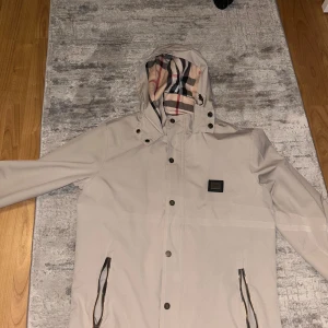 Beige vindjacka från Burberry - Snygg beige vindjacka från Burberry med klassiskt rutigt foder i huvan och insidan. Jackan har huva, dragkedja och knappar framtill samt två fickor med dragkedja. Perfekt för blåsiga dagar och stilren look.