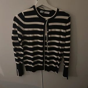 Svartvit randig kofta från Zara - Säljer en svart och vit randig kofta från Zara med rund halsringning och pärlknappar framtill. Koftan har långa ärmar och är tillverkad i ett mjukt stickat material. Perfekt för lager-på-lager och enkel att styla.
