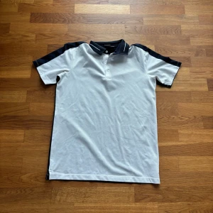 Vit och blå pikétröja med krage - Säljer en stilren pikétröja i vitt med blå detaljer på axlar och krage. Tröjan har korta ärmar, knappar vid halsen och klassisk pikékrage. Perfekt för en sportig och avslappnad look.