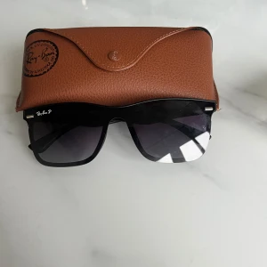 Svarta solglasögon från Ray-Ban - Snygga svarta solglasögon från Ray-Ban med klassisk fyrkantig form och mörka tonade glas. Kommer med ett brunt fodral i läderimitation. Perfekta för soliga dagar och ger en cool look.