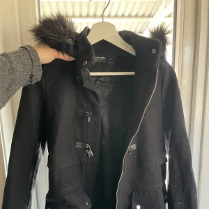 Svart kappa med fuskpäls från Zara  - Säljer en svart kappa från Zara med huva och fuskpälsdetalj. Jackan har dragkedja, knappar och stora fickor framtill. Storlek XS