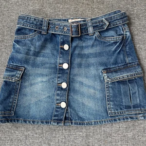 Jeanskjol med fickor och bälte från Next - Säljer en blå jeanskjol från Next med stora fickor på sidorna, bälte i midjan och knappar framtill. Knappt använd 