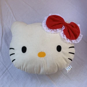 Hello Kitty diadem med röd rosett - Supergulligt diadem inspirerat av Hello Kitty, med en stor röd rosett dekorerad med vit spets. Diademet är i mjuk, vit plysch och har klassiska Hello Kitty-detaljer som svarta ögon och morrhår samt gul nos.