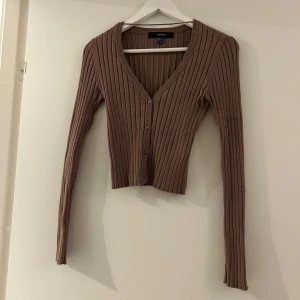 Brun ribbad kofta - Säljer en brun ribbad kofta från Vero Moda med v-ringning och knappar framtill. Modellen är croppad.